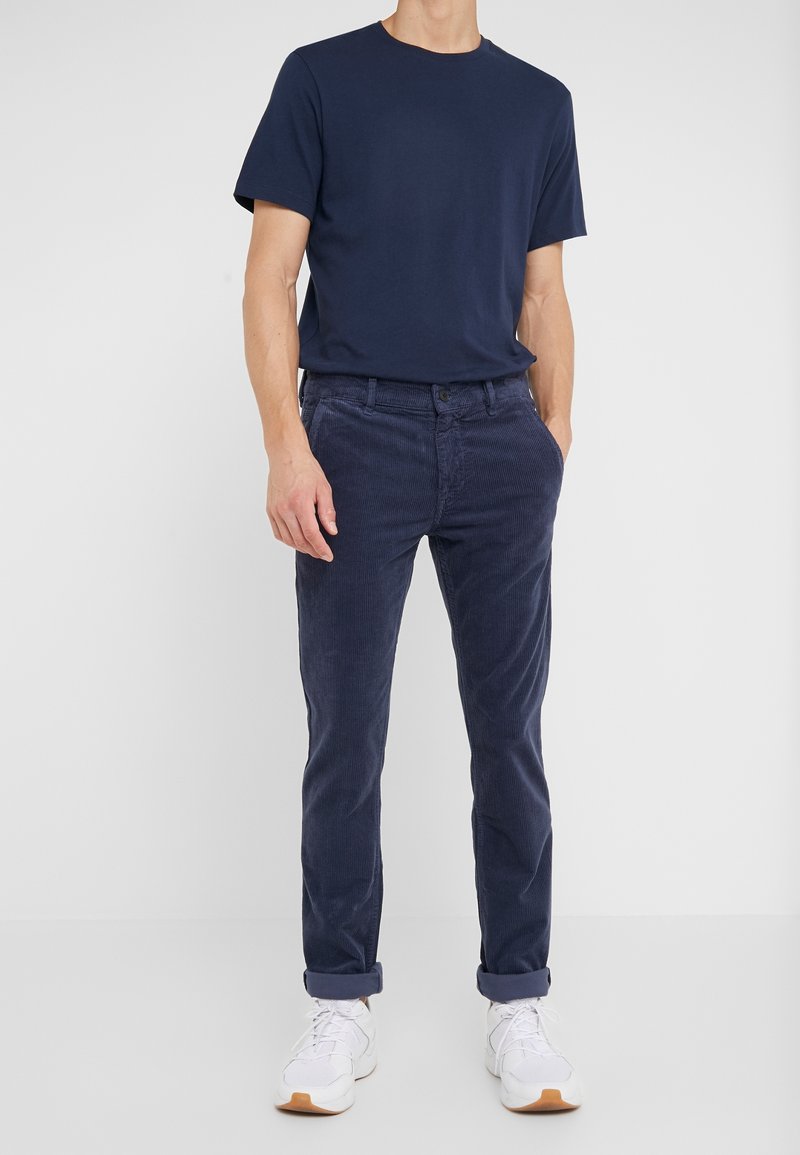 Laivastonsininen puuvillainen t-paita yhdistettynä tummansinisiin corduroy-housuihin, jotka on kääräisty lahkeista. Valkoiset urheilukengät täydentävät asun.