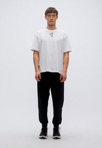 Άσπρο βαμβακερό μπλουζάκι με στρογγυλή λαιμόκοψη και κοντά μανίκια, με μαύρη γραφική λεπτομέρεια στο κέντρο. Συνδυάζεται με μαύρο παντελόνι jogger.