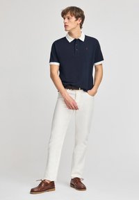 Marineblaues Polohemd mit weißem Kragen und Manschetten, cremefarbene Hose und braune Schuhe; ausgestattet mit Knopfverschluss und rotem Logo auf der Brust.