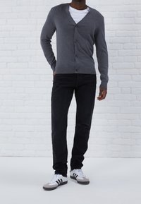 Cárdigan gris con escote en V, botones en la parte delantera y puños acanalados, combinado con jeans negros y zapatillas blancas con acentos grises. Fondo de ladrillo blanco.