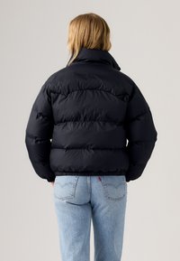 Chaqueta acolchada en azul marino oscuro, con un alto cuello, secciones acolchadas y puños elásticos. Llevada con jeans de un azul claro. Textura suave.
