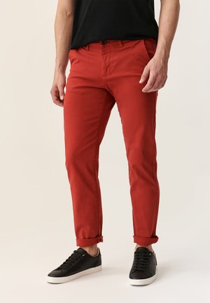 Rote Baumwoll-Chinos im Slim-Fit-Stil, mit aufgerollten Säumen und Seitentaschen, kombiniert mit schwarzen Schnürschuhen auf einem neutralen Hintergrund.