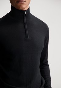 Pullover nero a maniche lunghe con colletto alto e zip a un quarto. Texture liscia con un design aderente e dettagli minimi.