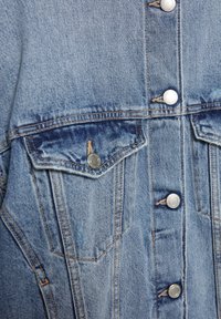 Jeans jakna v svetlo modri barvi. Ima srebrne gumbe, dve sprednji žepi in rahlo zbledelo teksturo, ki poudarja podrobnosti šivanja.