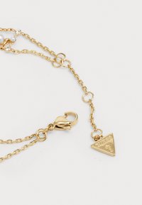 Gouden ketting met een schakeldesign, voorzien van een kreeftsluiting en een driehoekige hanger met het opschrift "GUESS." Glad, glanzend oppervlak.
