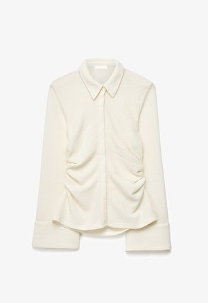 Ilgas rankovėmis kreminės spalvos tekstūruotas marškinys su aštriu apykakliu, priekiniais mygtukais ir sutrauktomis audinio detalėmis kiekvienoje liemens pusėje.