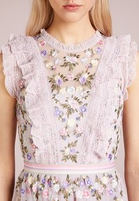 Woman wearing a sheer lavender dress with multicolored floral embroidery, ruffled lace sleeves, and a pastel striped waistband.
