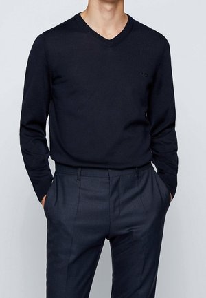 Mand iført en sort V-hals sweater og mørkegrå ternede habitbukser med hænderne i lommerne mod en ensfarvet baggrund.