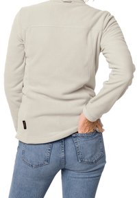 Ljusbeige fleecejacka med långa ärmar, med rundad hamn och en liten logotagg längst ner till vänster. Buren över blå jeans.