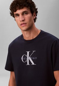 Tmavomodré bavlnené tričko s vycentrovaným bielym potlačeným logom "Calvin Klein" so štýlizovaným sivým nápisom "CK". Okrúhly výstrih, krátke rukávy.