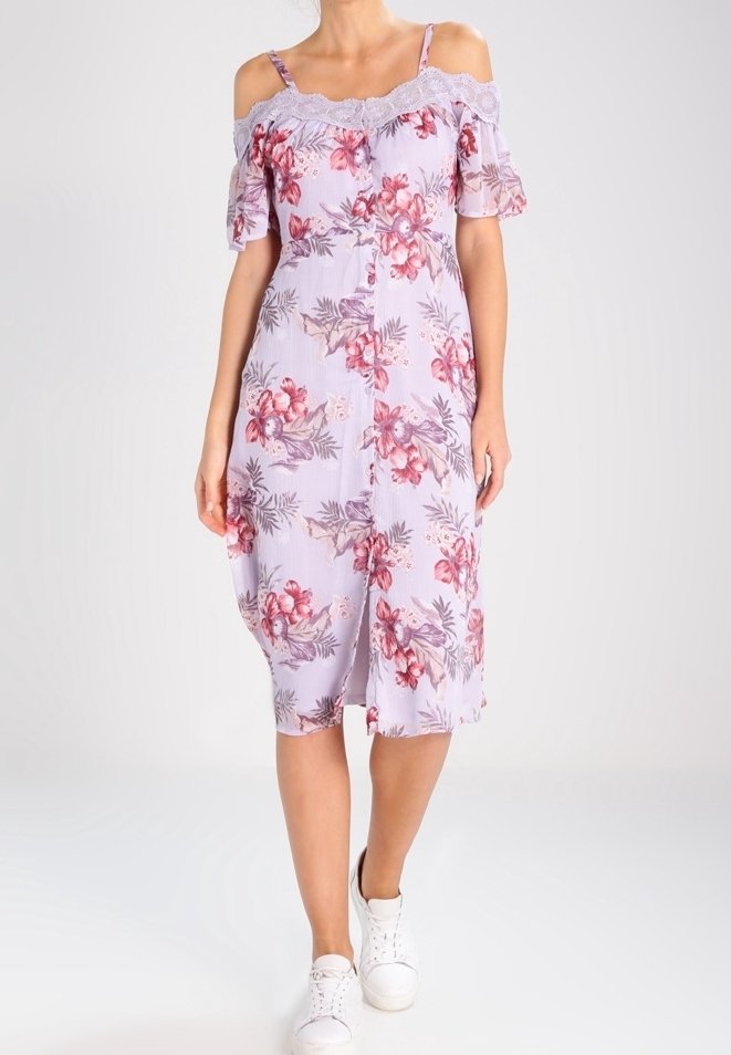 Femme portant une robe midi lilas à épaules dénudées avec un motif floral rose et des baskets blanches, debout devant un fond uni.