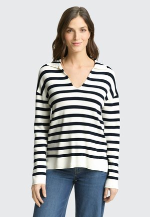 Femme aux cheveux bruns mi-longs portant un pull à col en V à rayures horizontales blanches et noires et un jean bleu, debout devant un fond uni.