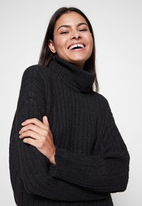 Schwarzer gerippter Rollkragenpullover mit langen Ärmeln, der eine grobe Stricktextur und eine lässige Passform aufweist. Modell lächelt mit verschränkten Armen.