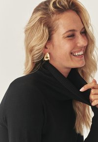 Eine lächelnde Frau mit blonden Haaren trägt einen schwarzen Rollkragenpullover und goldene ovale Ohrringe, während sie den Kragen des Pullovers mit ihrer Hand hält.