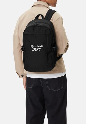 Osoba nesoucí černý batoh Reebok přes béžovou bundu a tmavé kalhoty, otočená zády k jednobarevnému světlému pozadí.