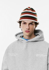 Grijze capuchontrui met witte tekst; blauw T-shirt eronder. Gebreide muts met oranje, groene en witte strepen. Neutrale achtergrond.