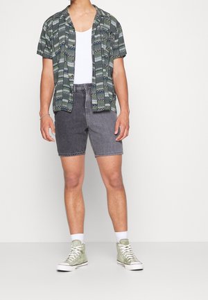 Camicia a maniche corte con motivo verde e nero, abbinata a pantaloni corti in denim grigi. La calzatura include sneakers alte verde chiaro.