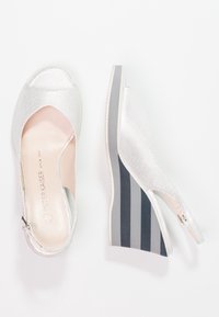 Chaussure compensée blanche perforée avec un design à bout ouvert et un talon compensé à rayures grises contrastantes. Dotée d'une fermeture éclair métallique sur le côté.