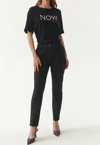 Schwarzes T-Shirt mit "NOW" in buntem Blumenmuster, kombiniert mit schwarzen engen Hosen und lila High Heels. Glatte Textur und lässiges Design.