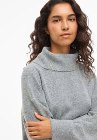 Grauer Strickpullover mit einem strukturierten Finish, locker sitzenden Ärmeln und einem weiten Kragen, getragen mit verschränkten Armen.