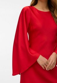 Sarkana kleita ar garām, zvana formas piedurknēm. Audumam ir gluda tekstūra, tam ir apaļa izgriezuma un pieguļošs siluets.