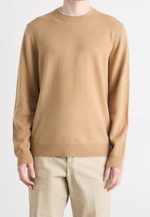 Beige Rundhals-Pullover aus weichem Material. Mit gerippten Bündchen und Saum, sowie einem kleinen, tonal gehaltenen Logo auf der Vorderseite. Kombiniert mit hellen Hosen.
