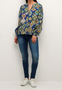 Kobieta ubrana w niebieską, kwiatową bluzkę z długim rękawem, dopasowane niebieskie dżinsy i białe trampki, stojąca na tle jasnej, jednolitej powierzchni.