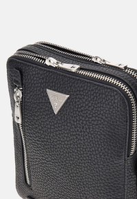 Schwarze, strukturierte Lederhandtasche mit silbernem Zubehör, doppelten Reißverschlüssen und einem dreieckigen Guess-Logo auf der Vorderseite. Kompaktes Design mit sichtbaren Nähdetails.