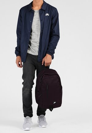 Personne portant une veste Nike bleu marine, un t-shirt gris, un jean noir, des baskets grises, tenant un sac à dos Nike noir par la poignée supérieure.