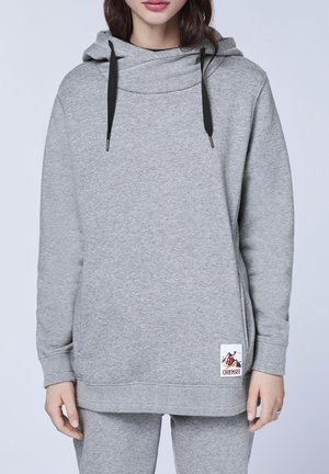 Vrouw draagt een lichtgrijze capuchontrui met zwarte trekkoorden en een klein logoplaatje bij de zoom, gecombineerd met bijpassende grijze broek.