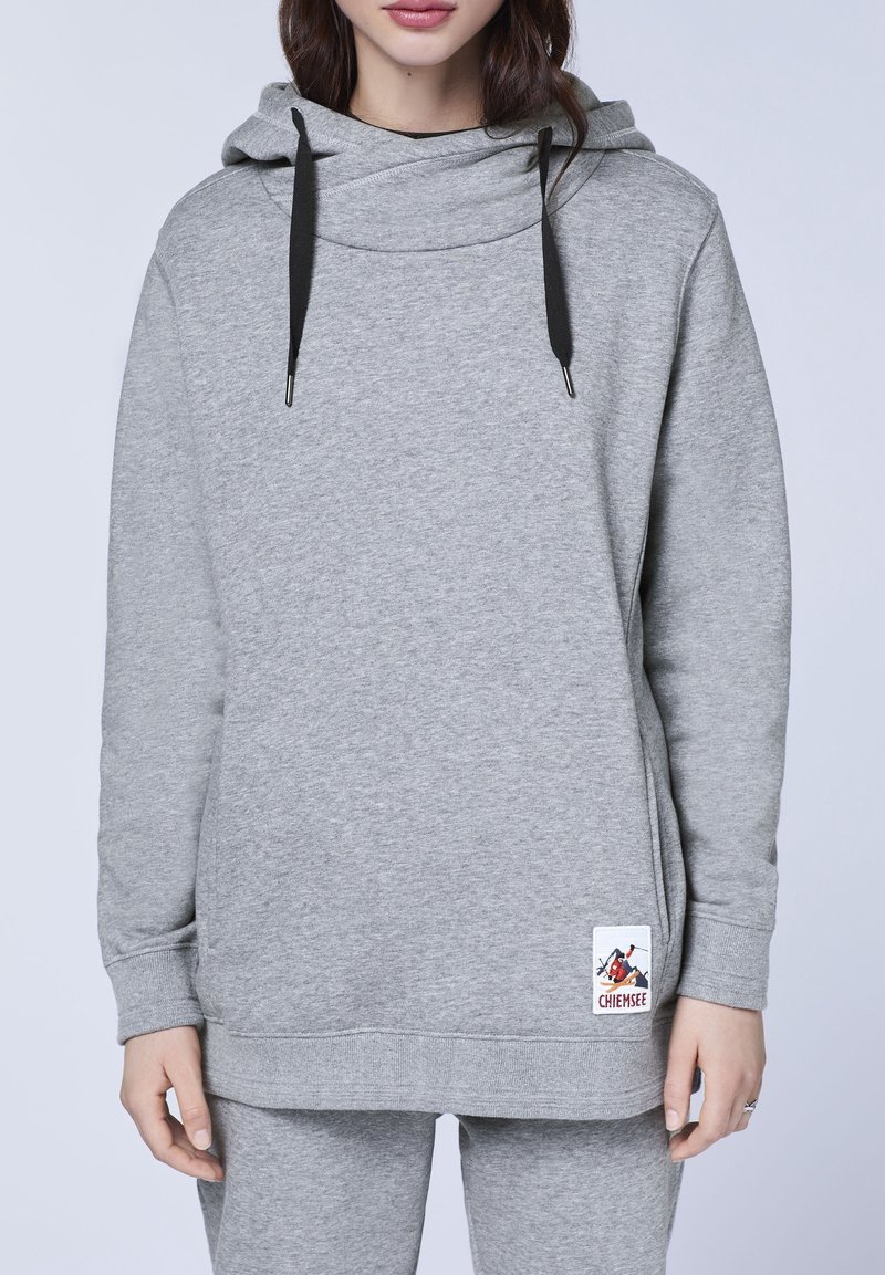 Femme portant un sweat à capuche gris clair avec des cordons de serrage noirs et un petit patch logo près de l'ourlet, assorti à un pantalon gris.
