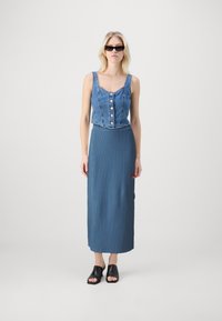 Un crop top en denim avec des boutons, associé à une jupe longue bleu côtelée. Des mules noires à bout ouvert complètent la tenue.