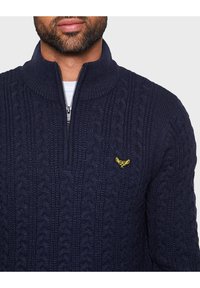 Marineblaue Strickpullover mit hohem Kragen und halbem Reißverschluss. Verfügt über ein kleines gelbes Logo-Emblem auf der linken Brust. Strukturiertes Gewebe.