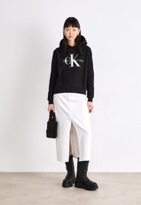 Juodas gobtuvas su baltu "CK" logotipu, baltos spalvos sijonas su priekiu plyšiu ir juodos spalvos kelių ilgio batai. Modelis laiko mažą juodą tekstūruotą maišelį.