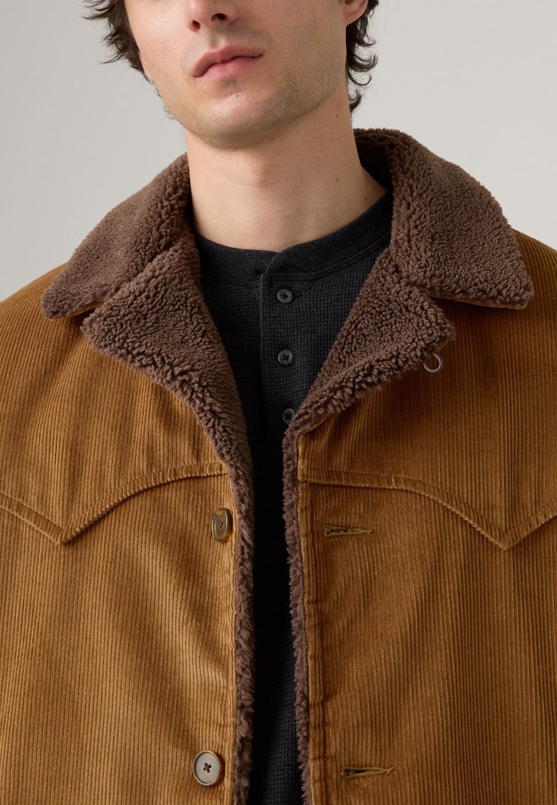 Chaqueta de pana marrón con un suave cuello de borreguito marrón, con cierres de botón y bolsillos en el pecho en diagonal. Llevada sobre una camiseta de textura oscura.