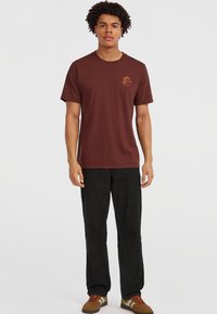 Bruine katoenen t-shirt met korte mouwen en een ronde halslijn. Heeft een klein oranje logo op de linkerborst. Draagt met zwarte broeken en bruine schoenen.