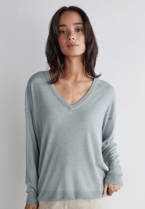 Ung kvinde med skulderlangt mørkt hår iført en lyseblå V-hals sweater og beige bukser foran en helt enkel væg.