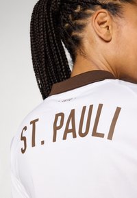 Hvit sportsdrakt med brun krage, med teksten "ST. PAULI" i fete, brune bokstaver på baksiden. Teksturert stoff synlig.