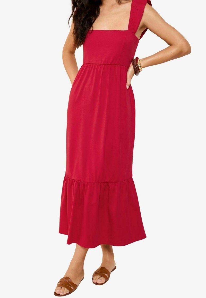 Femme portant une robe midi rouge sans manches avec un ourlet à volants, assortie à des sandales plates marron et des bracelets à un poignet.