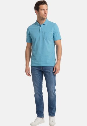 Mann steht und trägt ein hellblaues Poloshirt mit kurzen Ärmeln, blaue Jeans und weiße Sneakers, schaut zur Seite vor weißem Hintergrund.