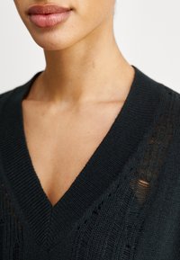 Svart stickad tröja med V-halsringning, texturerat mönster och öppna stygn detaljer längs halsen. Mjukt material med en jämn finish.