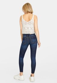 Top corto de encaje blanco con un patrón geométrico, combinado con jeans ajustados de color azul oscuro. La modelo lleva zapatillas blancas; la vista trasera muestra los detalles del diseño.