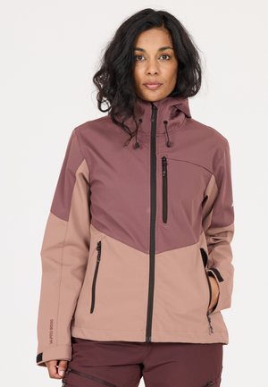 Vrouw met een medium huidskleur en donkergolvend haar draagt een tweekleurig mauve en roze jack met rits en meerdere ritszakken, staand tegen een effen achtergrond.