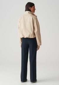 Beige cropped jack met een hoge kraag, elastische zoom en brede mouwen. Combinatie met donkere broeken, van achteraf gezien tegen een neutrale achtergrond.