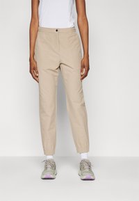 Pantalon beige à coupe décontractée, avec une fermeture à bouton à l'avant, des poches latérales et des poignets élastiqués, associé à des baskets grises.