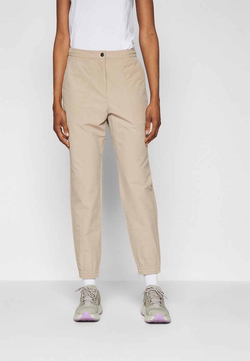 Pantalon beige à coupe décontractée, avec une fermeture à bouton à l'avant, des poches latérales et des poignets élastiqués, associé à des baskets grises.