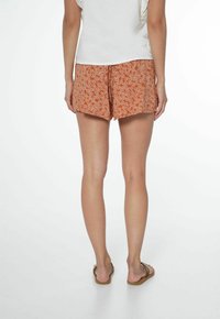 Oranžové šortky s bílými květinovými vzory, volného střihu a lemovanými nad středem stehna. V kombinaci s hnědými žabkami.