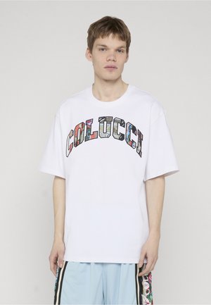 Young man standing wearing a white oversized T-shirt with colorful "COLUCCI" lettering and light blue shorts with side stripes.