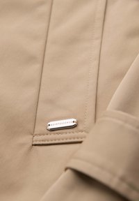 Gros plan sur un tissu beige montrant des coutures détaillées et une petite étiquette en métal argenté avec l'inscription « QUINTESSENCE ».