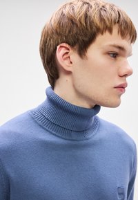 Suéter de gola alta azul claro em tecido malha macio. Apresenta um colarinho e punhos canelados, com um pequeno logo na zona do peito.